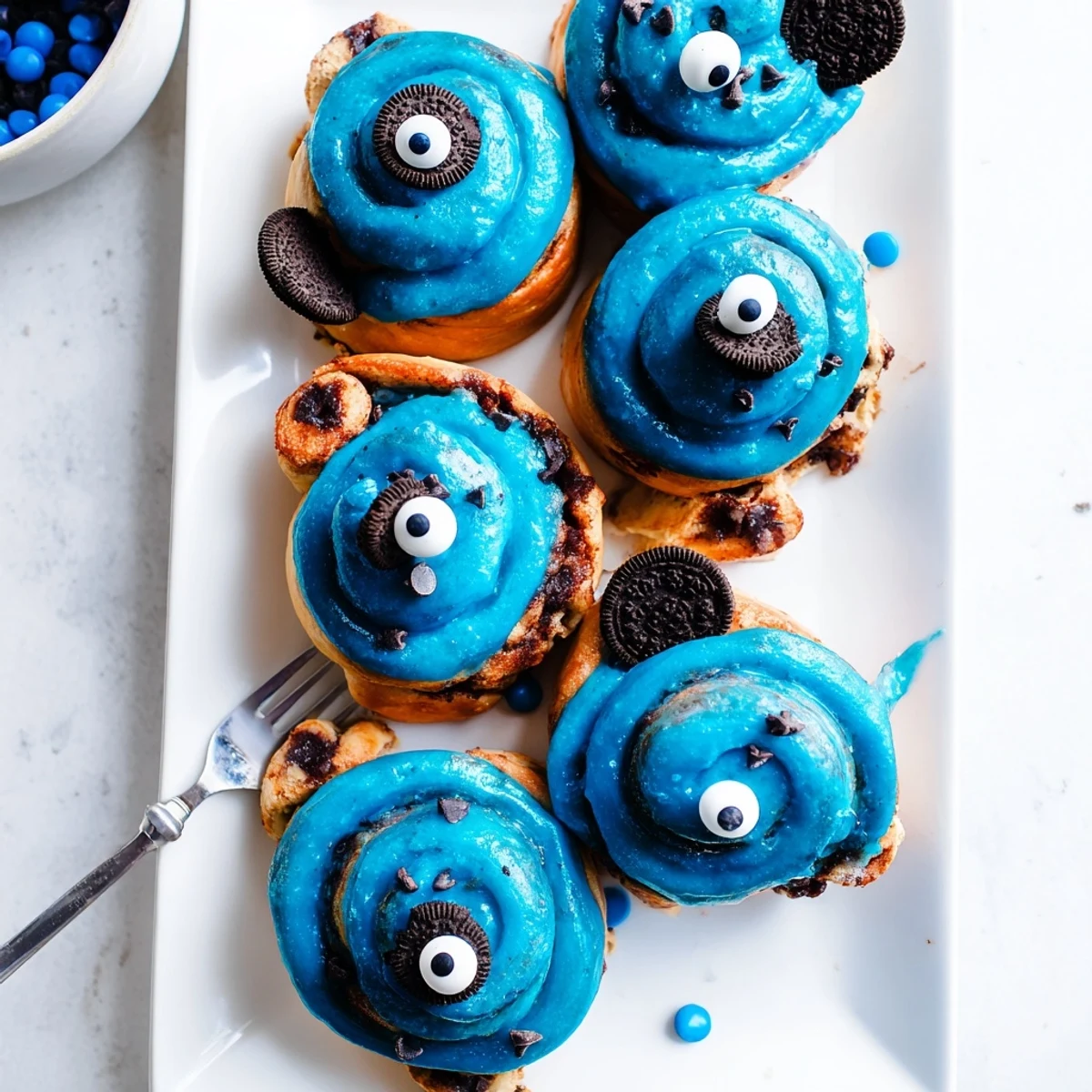 Warm blue cream cheese frosting generously drizzled over freshly baked Cookie Monster Cinnamon Rolls, with candy eyes staring back.